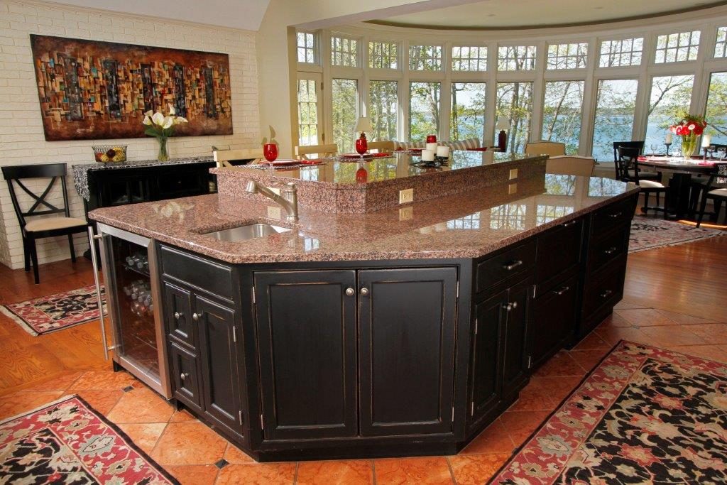 Spacious kitchen with a large granite island, dark cabinets, and bright windows overlooking trees and a lake.