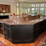 Spacious kitchen with a large granite island, dark cabinets, and bright windows overlooking trees and a lake.
