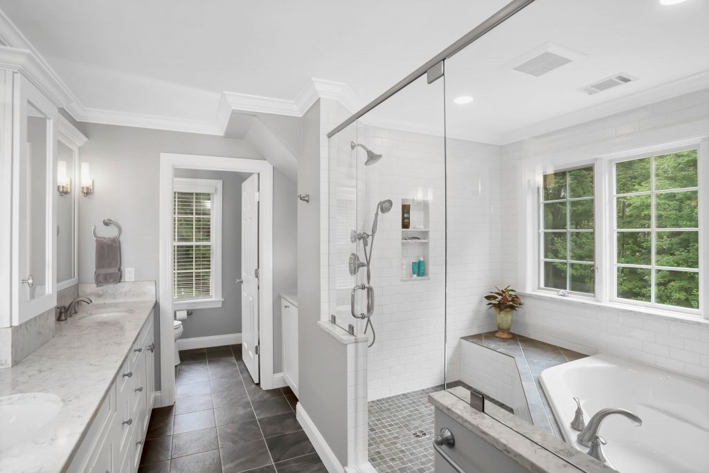 Modern bathroom with double sinks, a glass shower, large bathtub, white countertops, and a window with greenery outside.