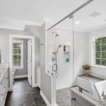 Modern bathroom with double sinks, a glass shower, large bathtub, white countertops, and a window with greenery outside.