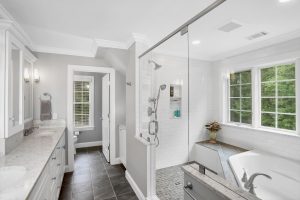 Modern bathroom with double sinks, a glass shower, large bathtub, white countertops, and a window with greenery outside.