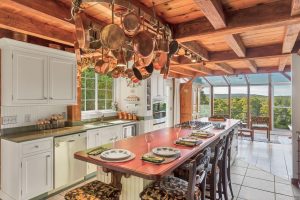Rustic kitchen with wooden beams, hanging copper pots, a long island, and large windows overlooking a scenic view.