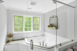 White bathroom with a corner bathtub, large window, potted plant, and wall decor with flowers above tiled surround.