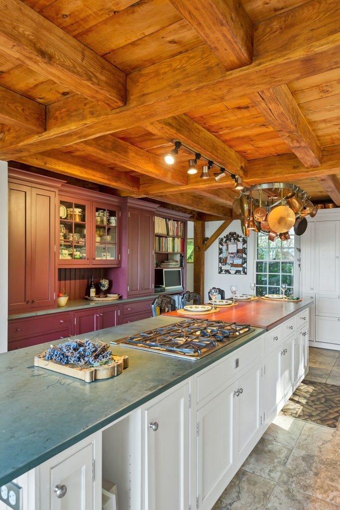 Rustic kitchen with wooden beams, white island, gas cooktop, hanging pots, and purple cabinetry in the background.