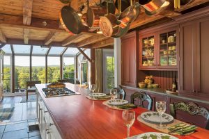 Sunny kitchen with a long wooden island, hanging pots, place settings, and large windows overlooking green scenery.