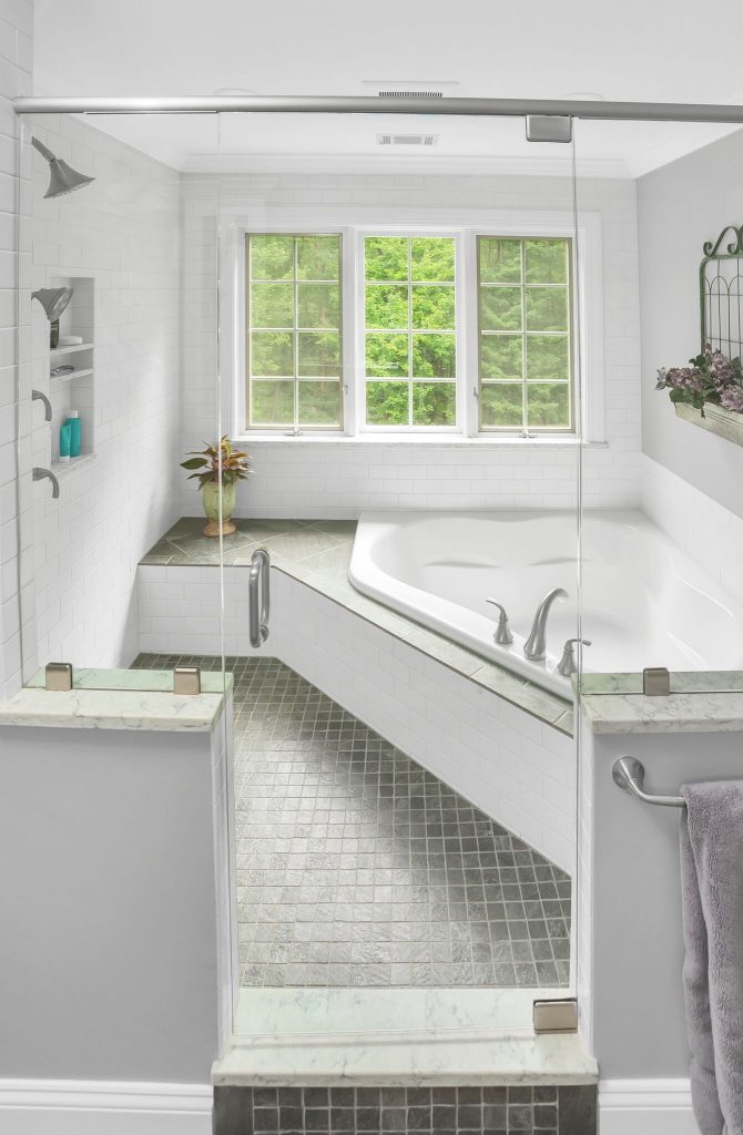 Modern bathroom with a large bathtub, glass shower door, tiled floor, and windows overlooking green trees.