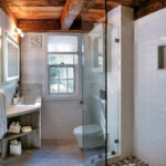 Modern bathroom with glass shower, wooden beams, white tile walls, single sink, and window letting in natural light.