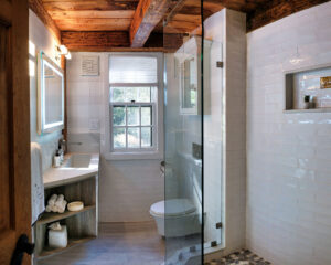 Modern bathroom with glass shower, wooden beams, white tile walls, single sink, and window letting in natural light.