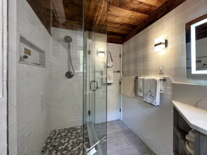 Modern bathroom with glass shower, wood ceiling, white tile walls, towel rack, and illuminated mirror above the sink.