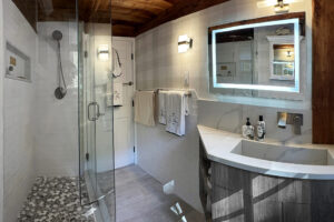 Modern bathroom with a walk-in shower, glass door, wooden ceiling, lighted mirror, and towel racks on white walls.