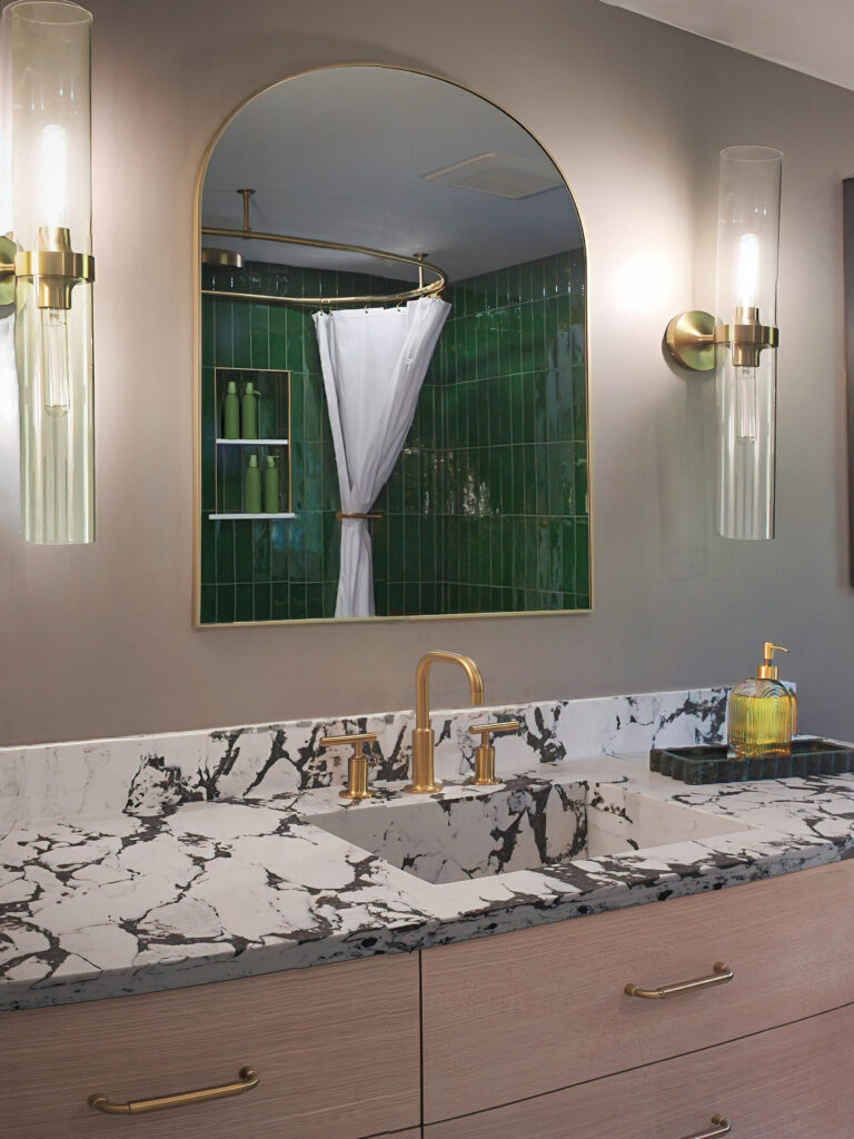 Modern bathroom vanity with marble countertop, gold fixtures, and a mirror reflecting a green-tiled shower area.
