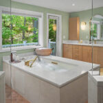 Modern bathroom with soaking tub, large windows, wood cabinets, double sinks, and views of green trees outside.