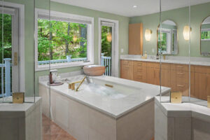Modern bathroom with soaking tub, large windows, wood cabinets, double sinks, and views of green trees outside.