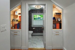 Bathroom with a window, flanked by two alcoves with shelves, books, and decor, set under a sloped ceiling.