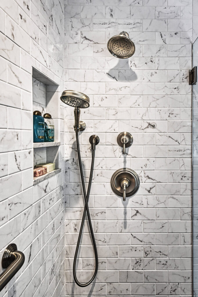 White tiled shower with two showerheads, a handheld wand, controls, and a shelf holding bottles and a bar of soap.