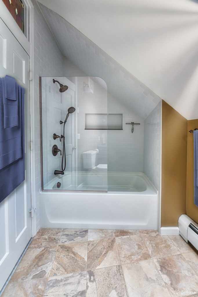 Modern bathroom with a bathtub-shower combo, glass partition, tiled walls, and sloped ceiling. Towels hang on nearby racks.