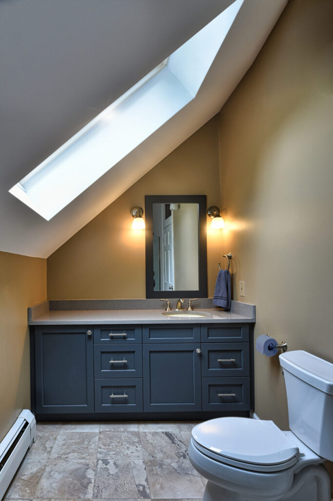 Attic bathroom with blue vanity, mirror, toilet, and skylight, featuring beige walls and tiled floor.