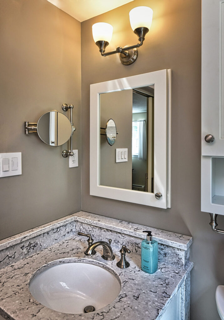 Small bathroom vanity with a marble countertop, sink, soap dispenser, mirror, and wall-mounted light fixture above.