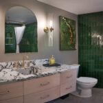 Modern bathroom with green tile walls, large mirror, marble countertop, and gold fixtures.