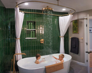 Two children play in a white bathtub with an overhead shower in a bathroom with green tile walls and gold fixtures.
