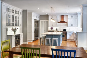 Modern kitchen with white cabinets, stainless appliances, island seating, and a wooden dining table with colorful chairs.