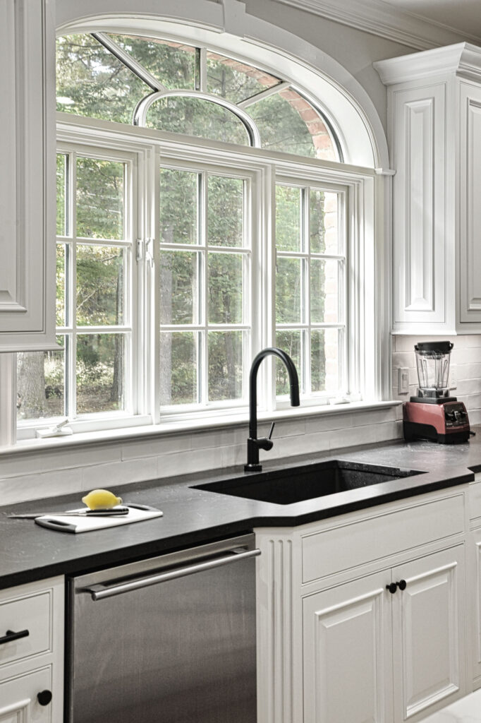 White kitchen with black sink and faucet, large window above, blender on counter, and a lemon on a cutting board.