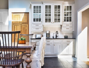 Bright kitchen with white cabinets, wooden table, green apples, and sunlight streaming through windows onto the floor.