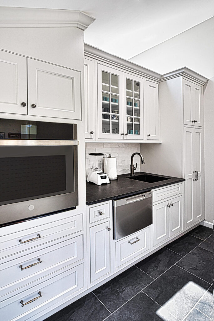 Modern white kitchen with black countertop, built-in oven, dishwasher, sink, blender, and upper glass cabinets.