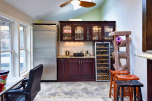 Home kitchenette area with dark cabinets, large fridge, wine cooler, cat tree, and stools on a tiled floor.