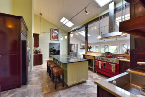 Modern kitchen with a large island, red stove, bar stools, skylights, and high ceilings with hanging lights.