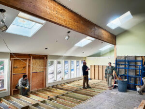 Three construction workers stand in a partially renovated room with exposed floor joists and large windows.