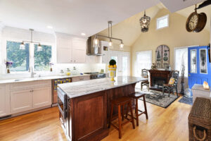 Bright kitchen with white cabinets, marble island, wooden stools, and a cozy sitting area near a blue door.