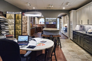 Kitchen design showroom with display cabinets, countertop samples, and a desk with a laptop and papers in the foreground.