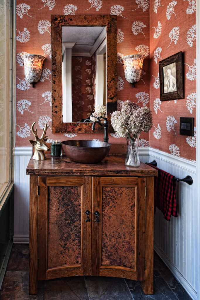 Rustic bathroom vanity with a round vessel sink, floral wallpaper, and wall sconces beside a large framed mirror.