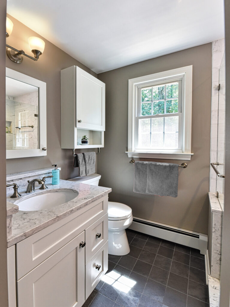 Small bathroom with white cabinets, gray walls, a window, marble countertop, and a towel hanging by the sink.