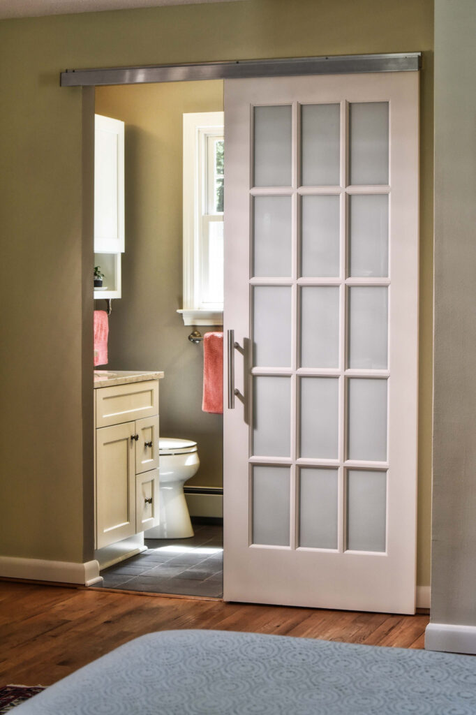 A white sliding glass door opens to reveal a bathroom with light cabinetry and pink towels hanging near the sink.