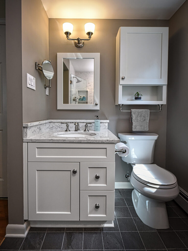 Small modern bathroom with gray walls, a white vanity, marble countertop, toilet, and upper cabinet above the toilet.