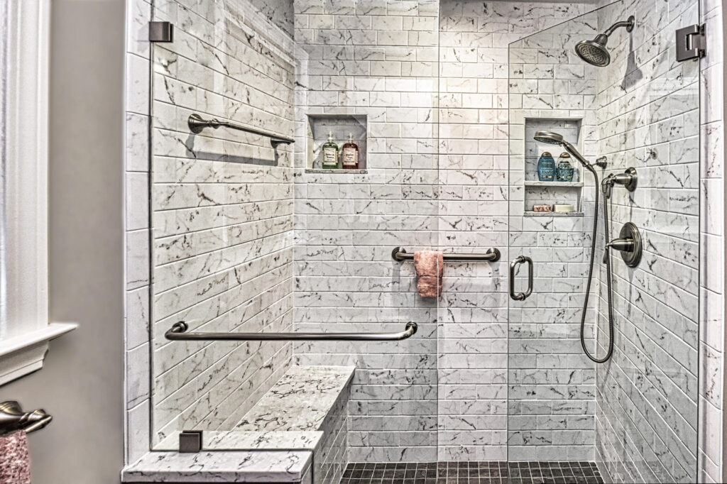 Modern walk-in shower with marble tile, built-in bench, grab bars, shelves, towel, and two showerheads.