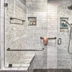 Modern walk-in shower with marble tile, built-in bench, grab bars, shelves, towel, and two showerheads.